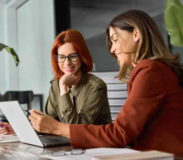 Two happy busy female employees working together using computer planning project. Middle aged professional business woman consulting teaching young employee looking at laptop sitting at desk in office